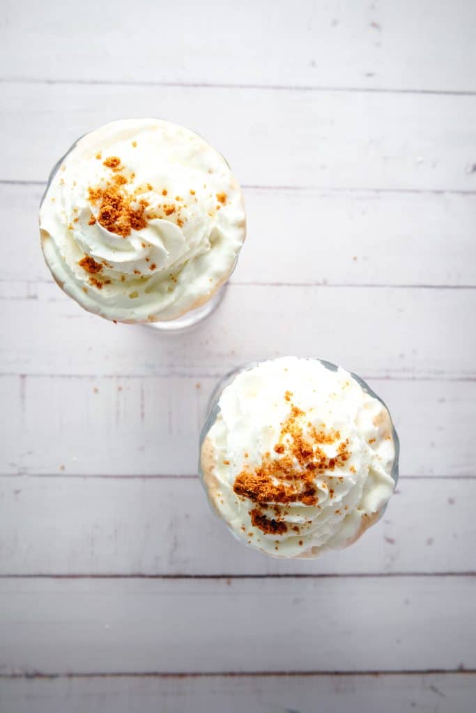 Two glasses with whipped cream and brown Biscoff crumbs on top, viewed from above on a white wooden surfaceโan irresistible Biscoff milkshake perfect for any sweet craving.