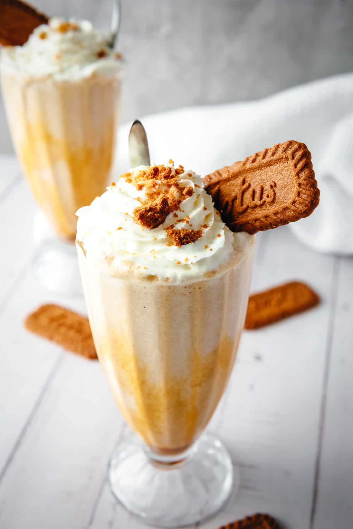 A Biscoff Milkshake topped with whipped cream, cookie crumbs, and a whole Biscoff cookie in a tall glass, served with a spoon.