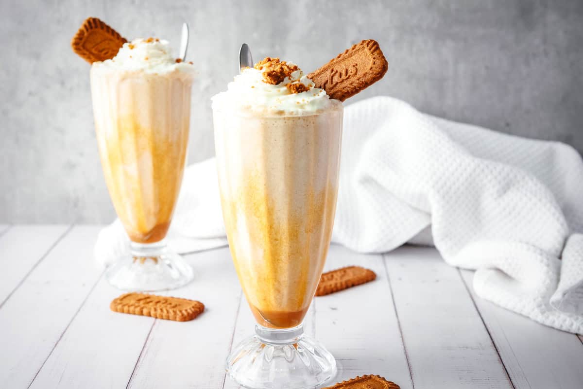 Two creamy Biscoff milkshakes in tall glasses topped with whipped cream and a cookie, on a white wooden surface.