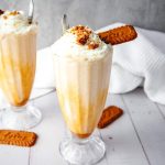 Two milkshakes with whipped cream and cookies in tall glasses on a white table with a white cloth, perfect for anyone craving a delicious Biscoff Shake.
