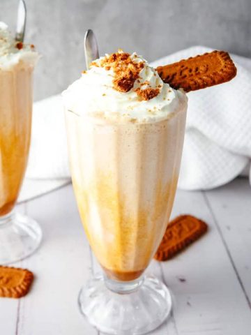 Two milkshakes with whipped cream and cookies in tall glasses on a white table with a white cloth, perfect for anyone craving a delicious Biscoff Shake.