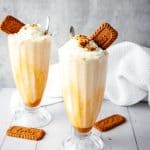 Two Biscoff milkshakes with whipped cream and cookies in tall glasses on a white table, with extra cookies nearby.