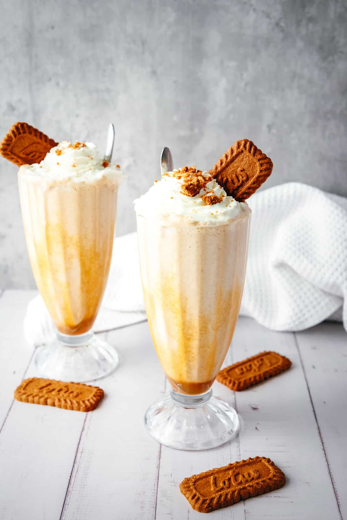 Two Biscoff milkshakes with whipped cream and cookies in tall glasses on a white table, with extra cookies nearby.