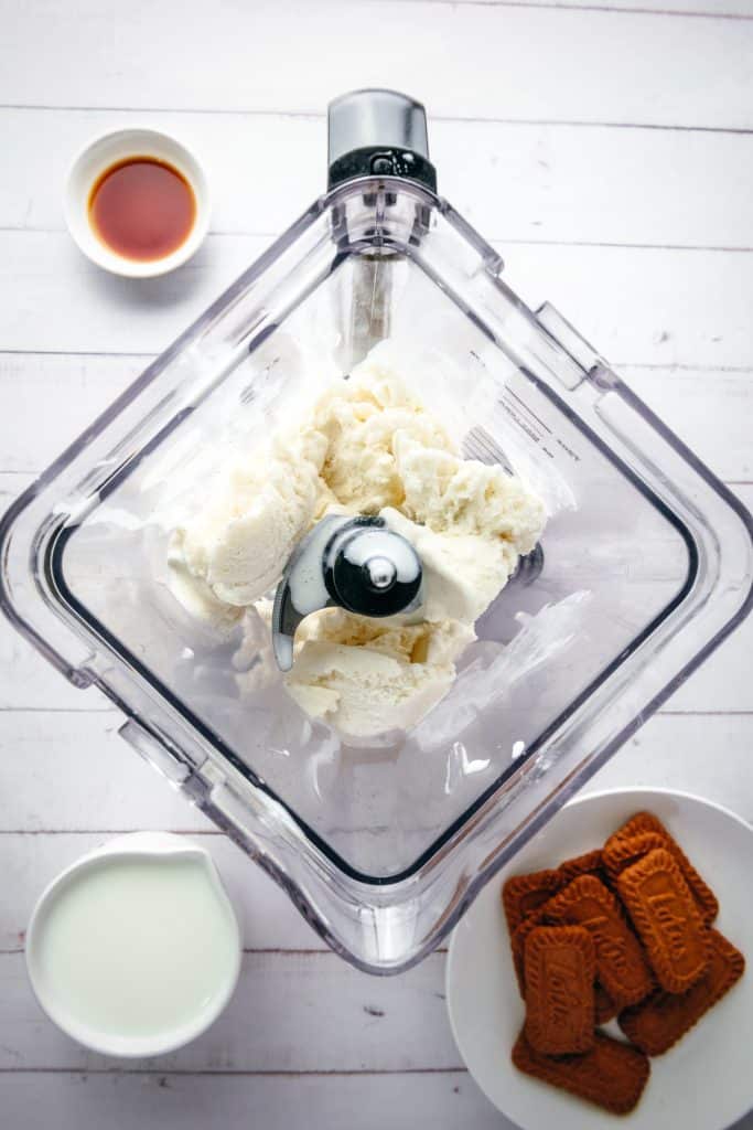 Vanilla ice cream in a blender with bowls of milk, cookies, and vanilla extract on a white table, perfect for creating a creamy Biscoff Milkshake.