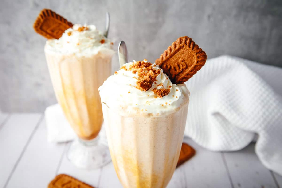 Two tall glasses of Biscoff milkshake topped with whipped cream and a cookie, with a white towel in the background.