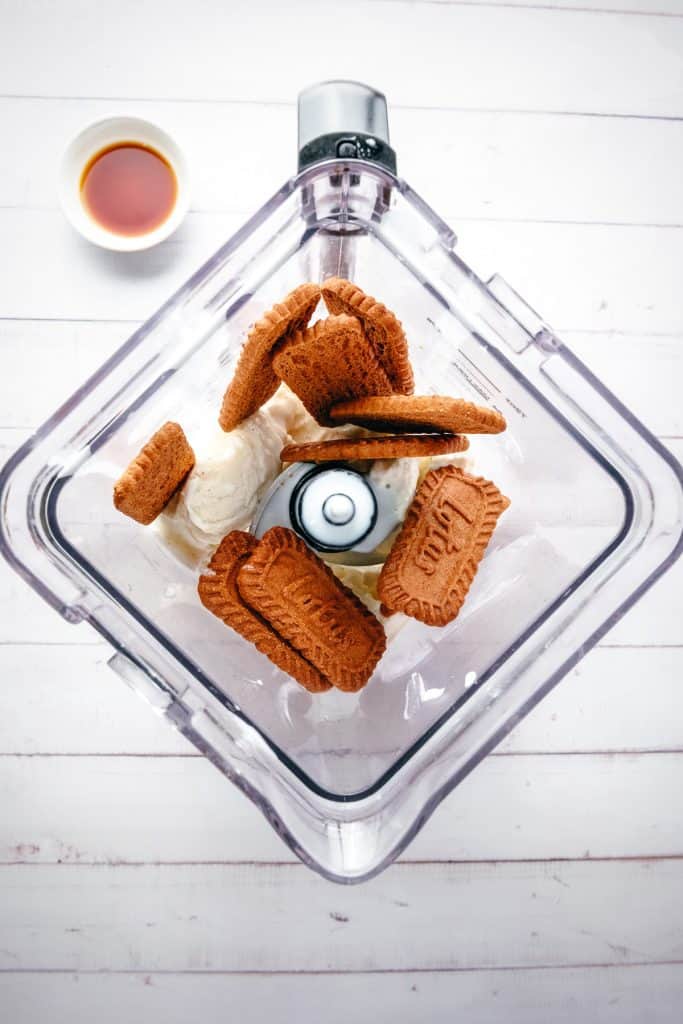 Top-down view of a blender with cookies and ice cream inside, ready to create a delicious Biscoff Milkshake, with a small bowl of liquid nearby on a white table.