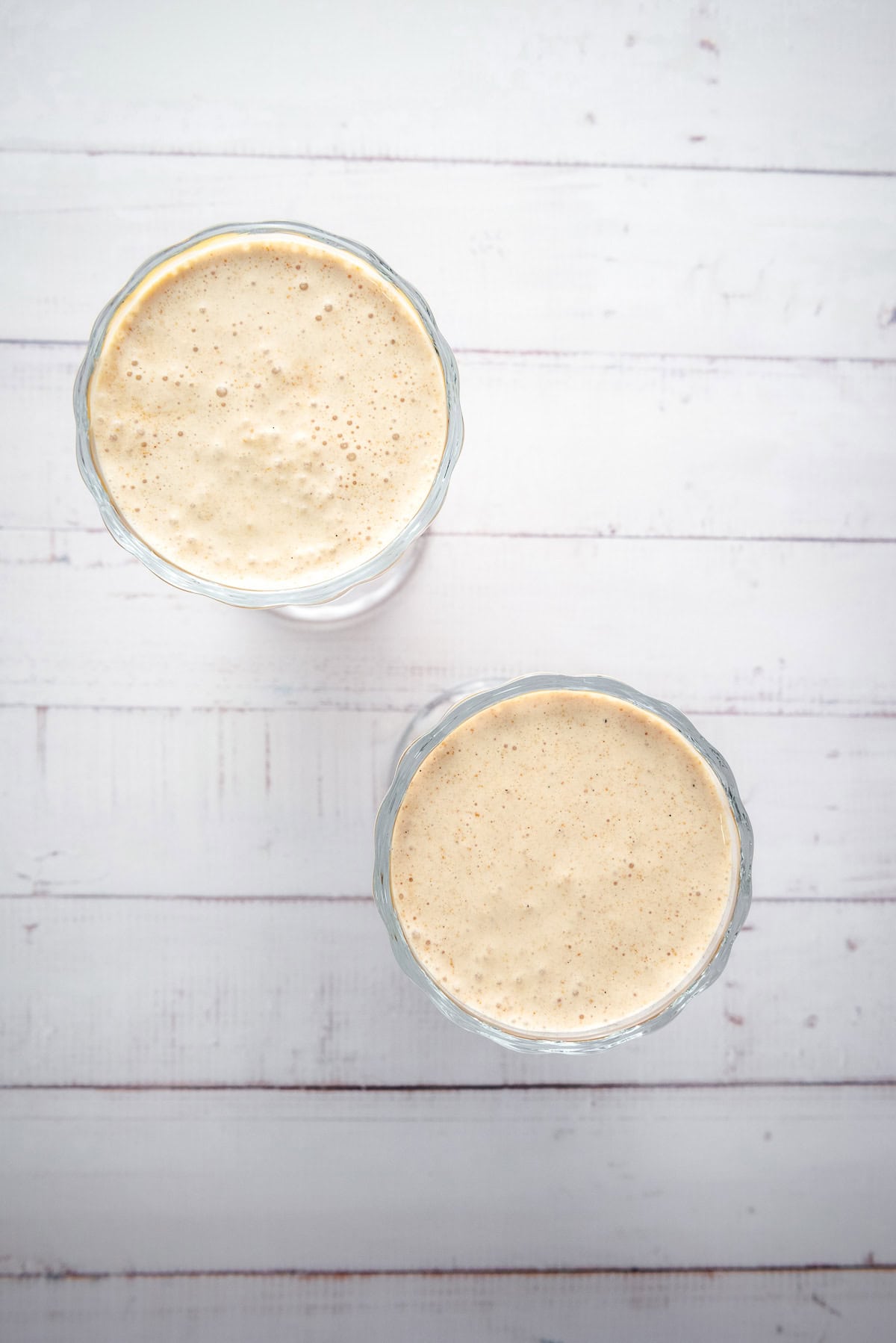 Two frothy, creamy Biscoff milkshakes in clear glasses, photographed from above on a white wooden surface.
