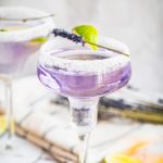 A vibrant lavender margarita mocktail in a sugar-rimmed glass, garnished with lime and sprigs of lavender, with fresh lime slices nearby.