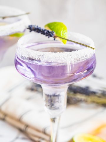 A vibrant lavender margarita mocktail in a sugar-rimmed glass, garnished with lime and sprigs of lavender, with fresh lime slices nearby.
