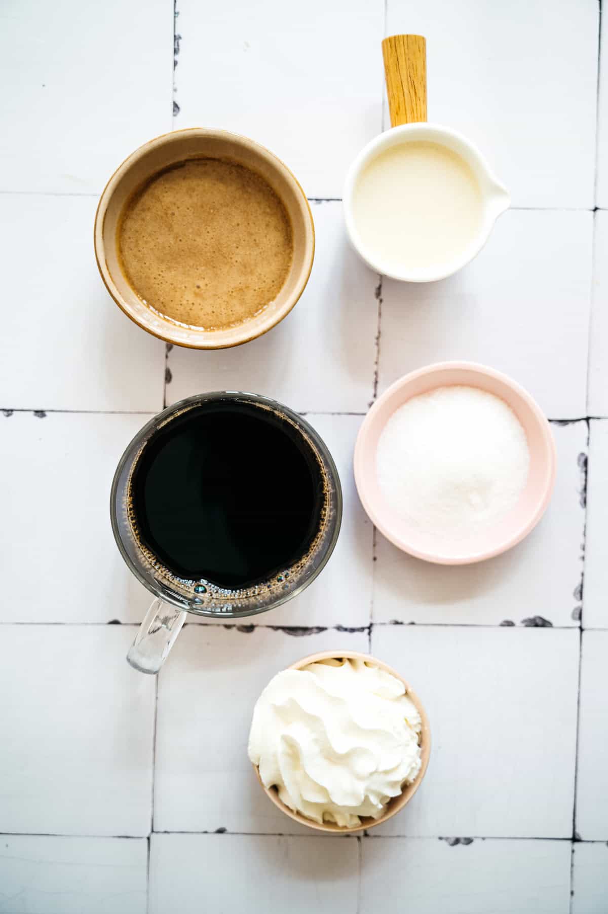 Five bowls and cups with coffee, sugar, cream, espresso, and whipped cream on a white tiled surface—perfect for crafting your own Oatmilk Frappuccino or caramel-inspired treat.