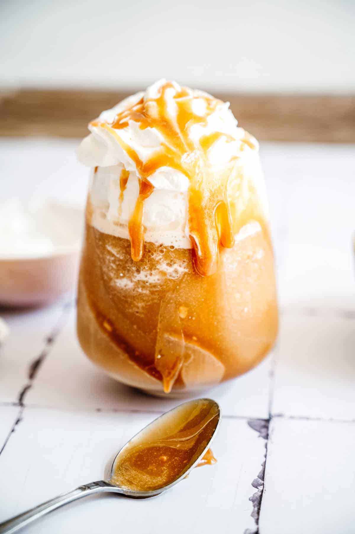Iced coffee topped with whipped cream and a drizzle of caramel sauce, featuring creamy oatmilk for a smooth finish—a delicious caramel frappuccino with a spoon of caramel sauce in front.