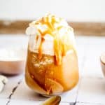 Iced caramel Frappuccino drink made with creamy oatmilk, topped with whipped cream and caramel drizzle, next to a spoon on a white surface.