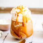 Glass of iced Caramel Frappuccino with whipped cream and caramel drizzle, next to a spoon on a white surface.