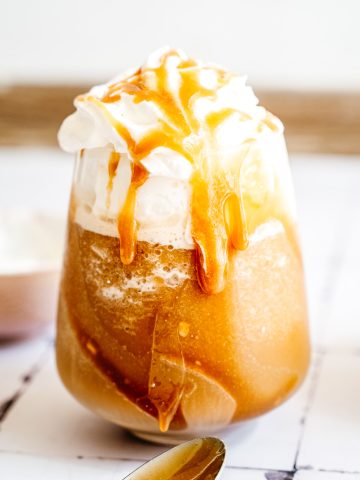 Glass of iced Caramel Frappuccino with whipped cream and caramel drizzle, next to a spoon on a white surface.