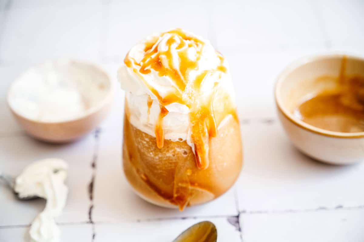 Iced coffee frappuccino topped with whipped cream and caramel sauce, surrounded by bowls and a spoon on a white surface.