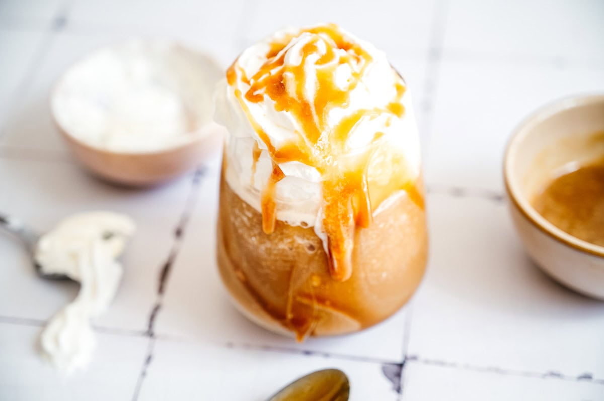 Iced oatmilk frappuccino topped with whipped cream and caramel sauce, with bowls and a spoon nearby on a white surface.