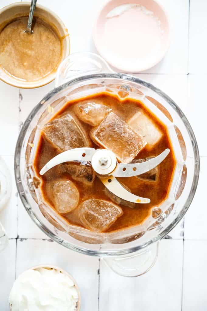 Overhead view of an oatmilk iced coffee Frappuccino in a blender with ice cubes and blender blades visible, surrounded by tempting toppings.