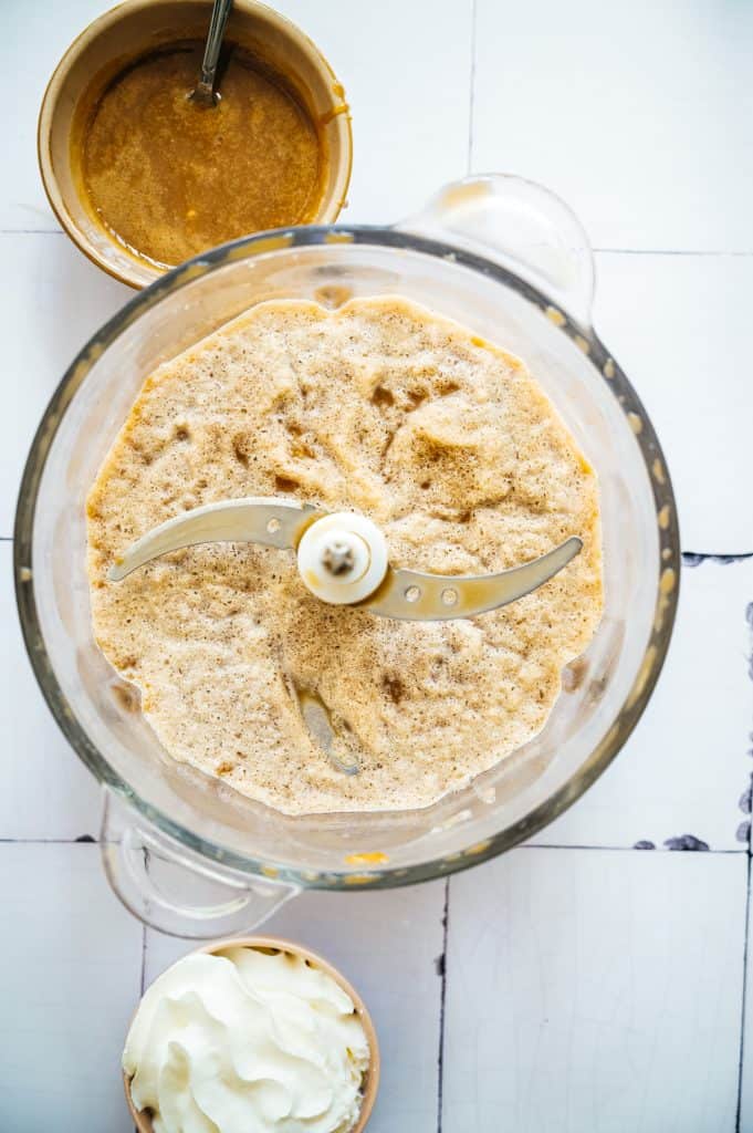 Blender with a frothy mixture, a bowl of whipped cream, and a bowl of caramel brown liquid on a white surface—perfect for crafting an oatmilk frappuccino.