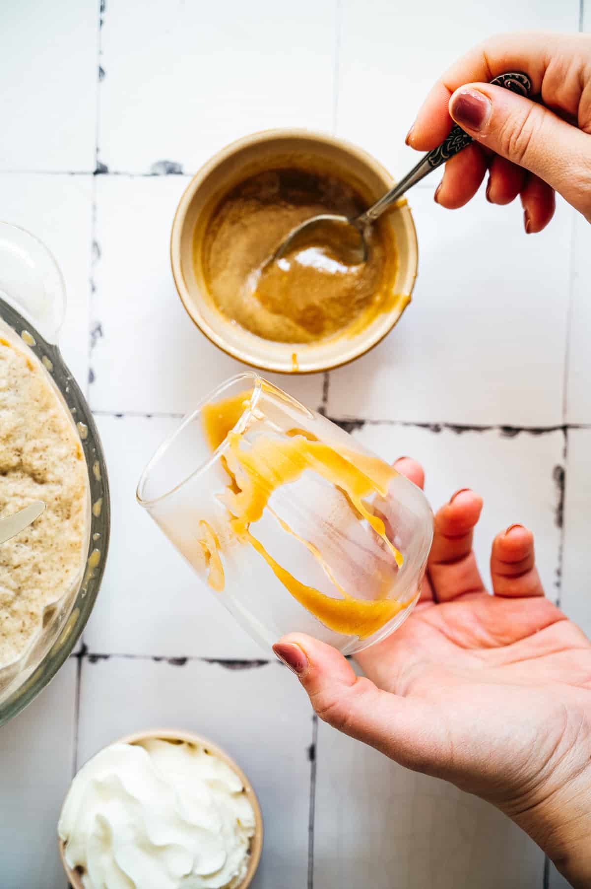 Hand drizzling caramel sauce inside a clear glass, with bowls of whipped cream and dessert nearby—perfect for creating a creamy Oatmilk Frappuccino or your favorite Caramel Frappuccino treat.