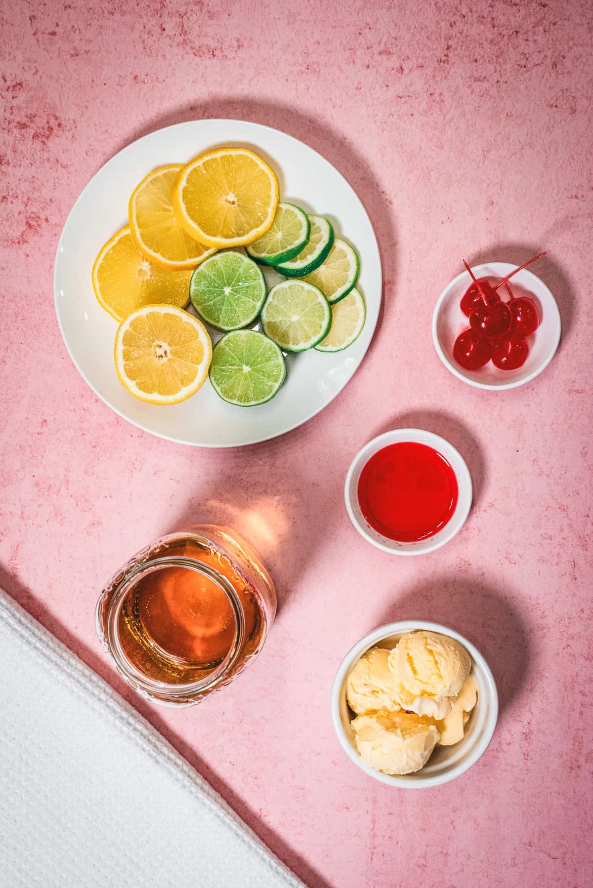 Sliced lemon, lime, cherries, syrup, vanilla ice cream, and a drink on a pink surface come together to create a delightful Shirley Temple Float.