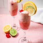 Two pink fizzy drinks in champagne glasses, garnished with lemon, lime slices, and cherries on a pink surfaceโperfect for enjoying as a light and refreshing Shirley Temple Float.