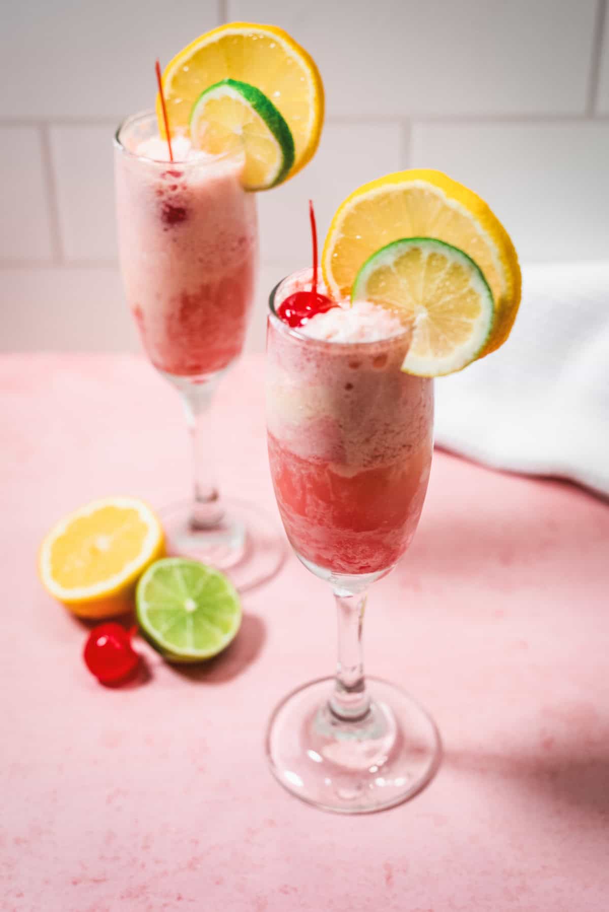 Two pink fizzy drinks in champagne glasses, garnished with lemon, lime slices, and cherries on a pink surfaceโperfect for enjoying as a light and refreshing Shirley Temple Float.