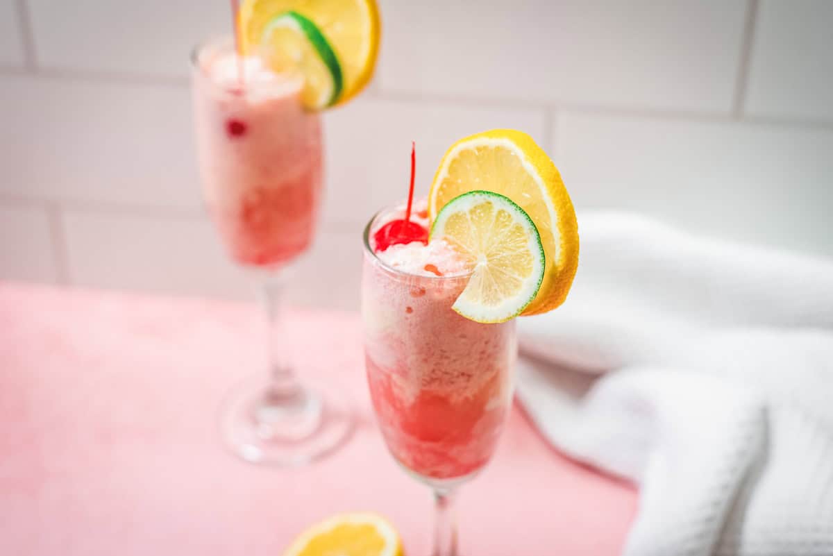 Two tall glasses of pink Shirley Temple float drinks topped with cherries, lemon, and lime slices sit on a pink surface with a white towel.