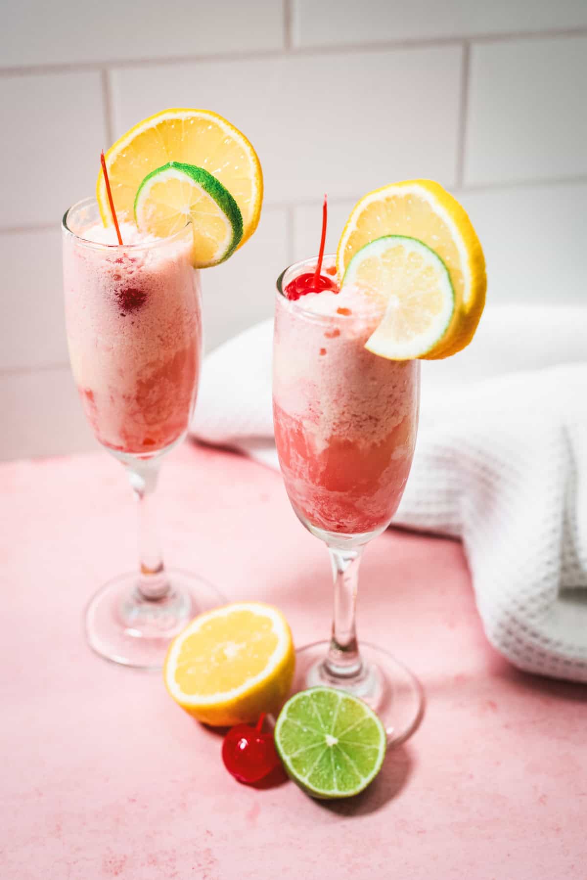 Two glasses of pink frozen Shirley Temple Float topped with lemon, lime, and cherries sit on a pink surface with fruit nearby.