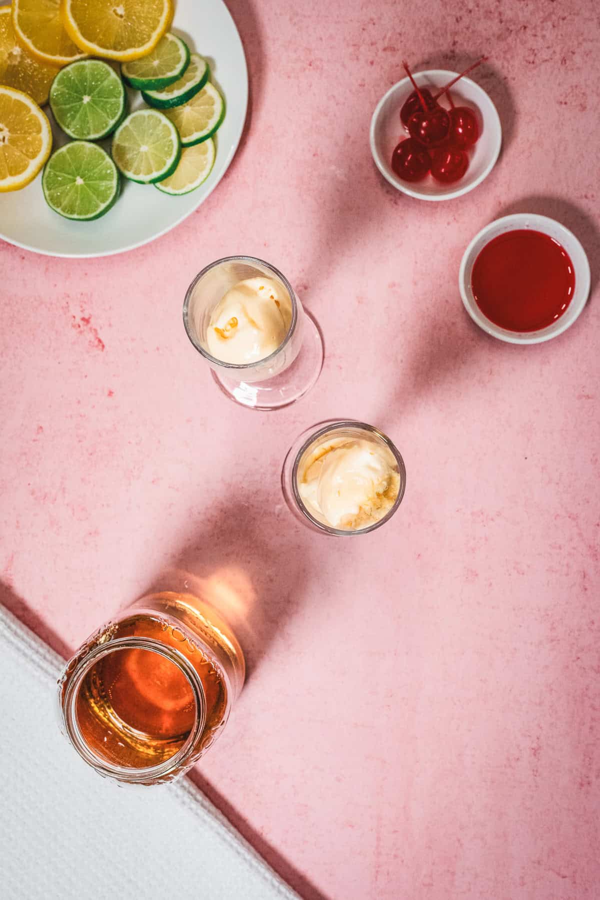Sliced citrus, cherries, and ice cream in glasses come together to create a delightful Shirley Temple Float, with a jar of liquid on a pink surface.