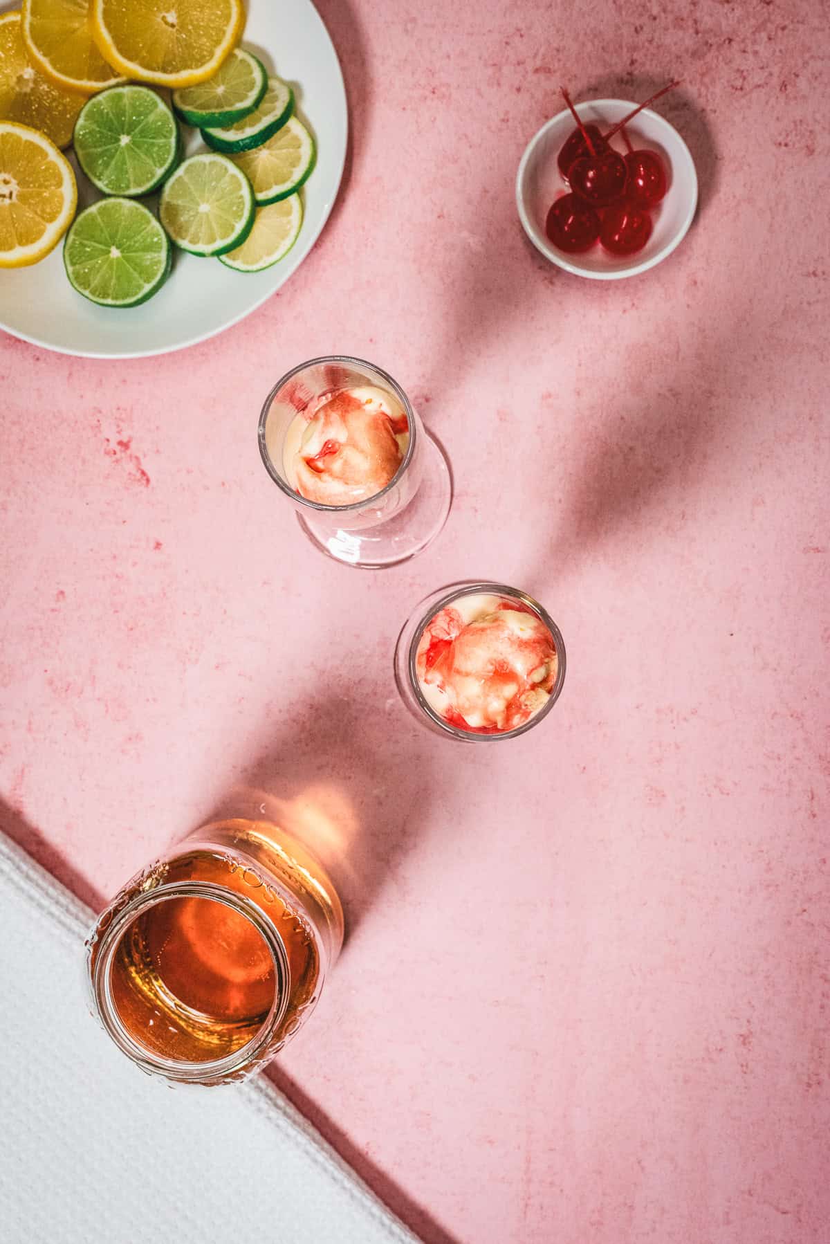 Three drinks with ice and cherriesโperfect for a refreshing Shirley Temple or even a delightful Shirley Temple Floatโsit beside a plate of sliced citrus and a small bowl of cherries on a pink surface.