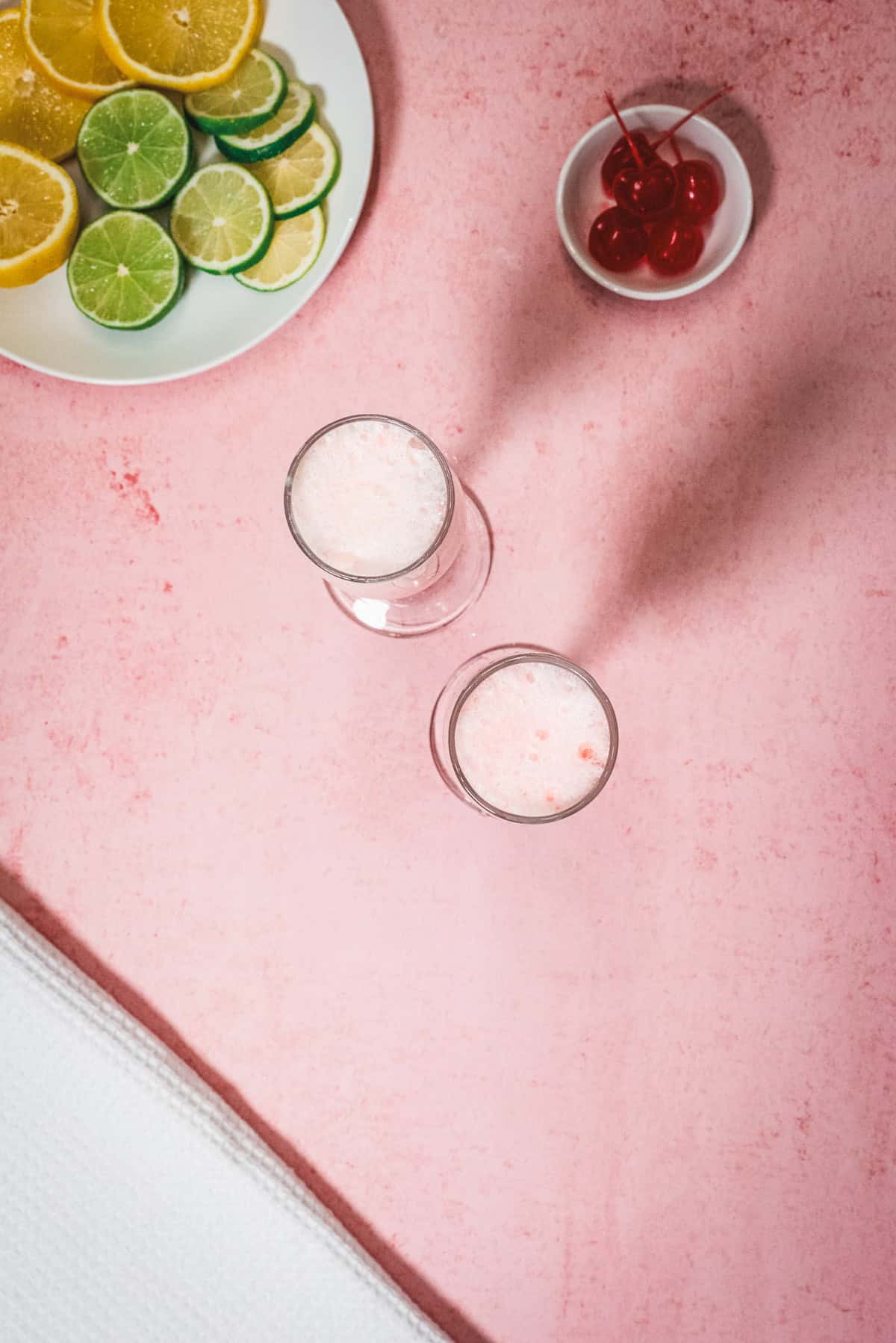 Two fizzy drinksโlike a classic Shirley Temple Floatโsit on a pink surface with sliced citrus fruits and cherries nearby.