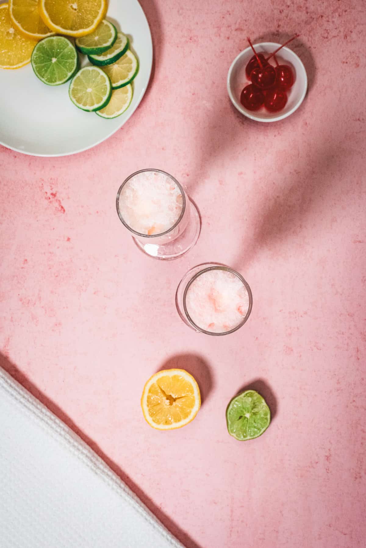 Two glasses with ice, lemon and lime slices, and cherries on a pink surface suggest a refreshing twistโperhaps a classic Shirley Temple or even a playful Shirley Temple Float. A plate and towel are visible nearby, adding to the inviting scene.