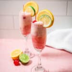 Two fruity cocktails inspired by the classic Shirley Temple float, with ice cream, orange, lemon, and cherry garnishes on a pink surface with citrus slices.