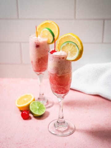 Two fruity cocktails inspired by the classic Shirley Temple float, with ice cream, orange, lemon, and cherry garnishes on a pink surface with citrus slices.