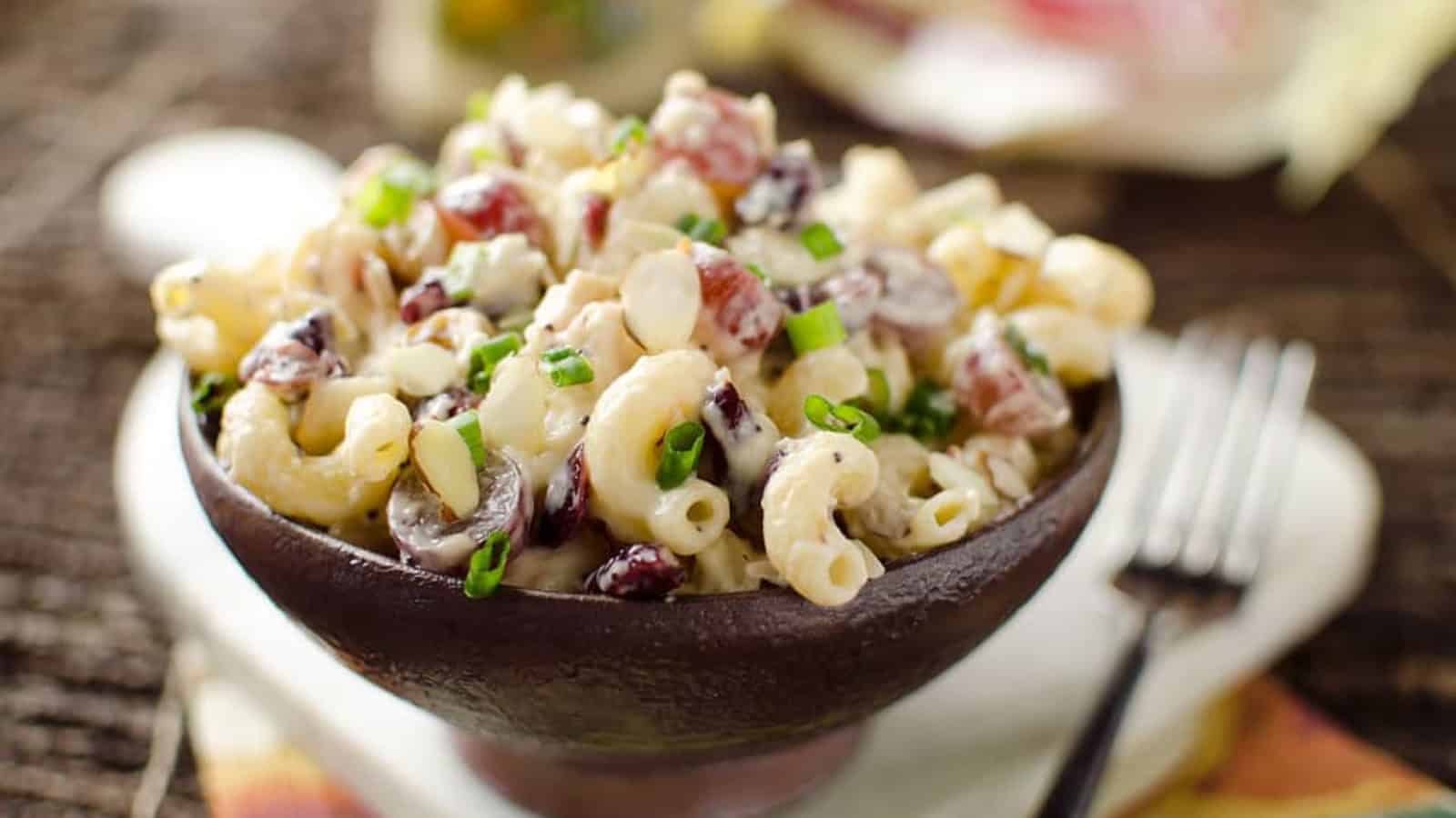 A bowl of macaroni salad with green onions, grapes, and creamy dressing sits next to a fork.