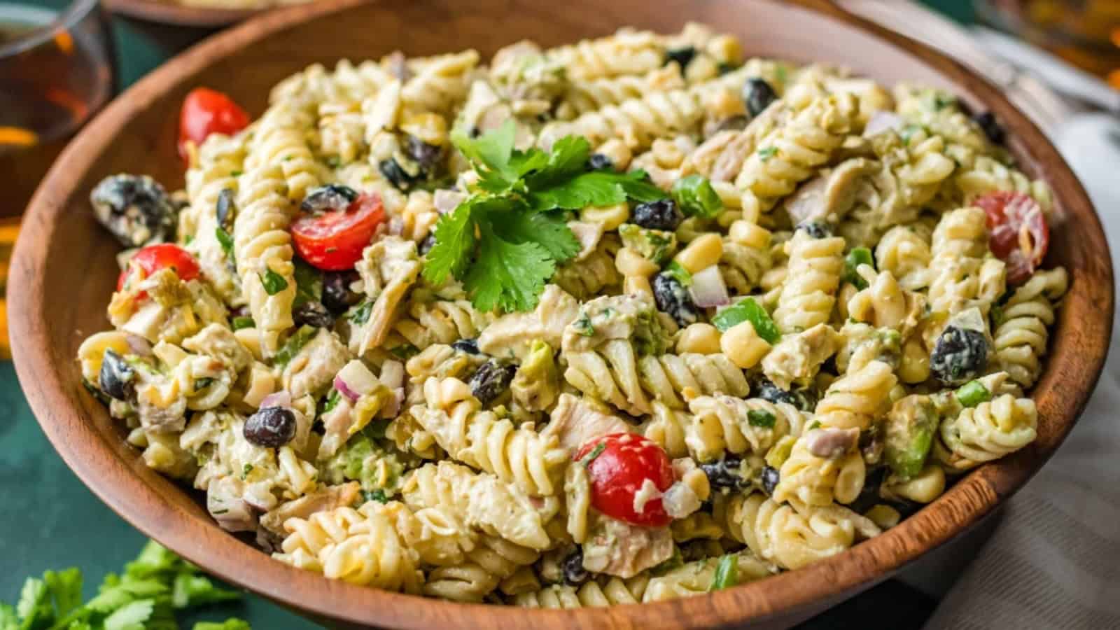 A wooden bowl filled with creamy pasta salad, cherry tomatoes, olives, corn, and fresh parsley on top.