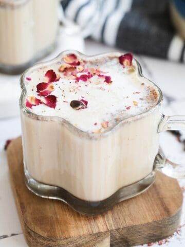 A glass mug of Chai Spiced Honey Latte with foamy milk and dried rose petals on top, placed on a small wooden board.