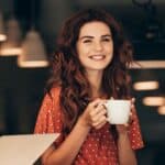 Smiling woman with long red hair holding a white mug in a cozy café, wearing a red patterned dress and savoring one of 7 coffee alternatives.