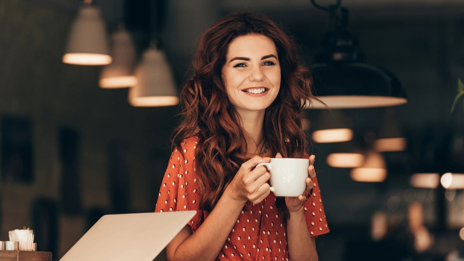 Smiling woman with long red hair holding a white mug in a cozy café, wearing a red patterned dress and savoring one of 7 coffee alternatives.
