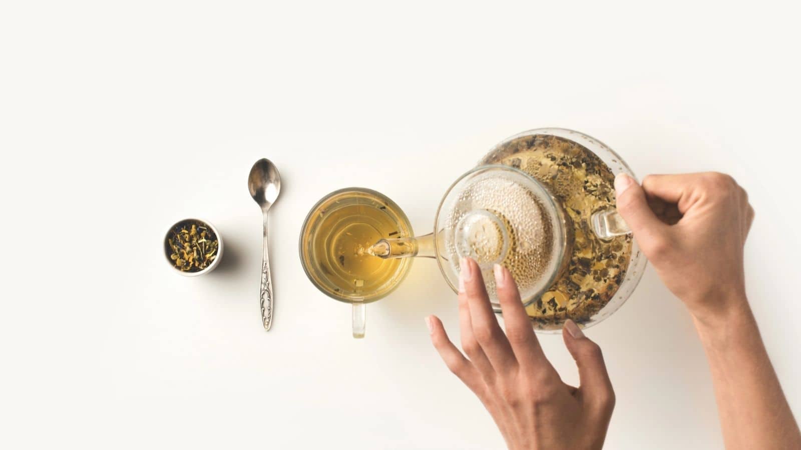 Hand pouring tea from a clear teapot into a cup, with a spoon and tea leaves on a white background, highlighting one of 7 coffee alternatives for those seeking a soothing drink.