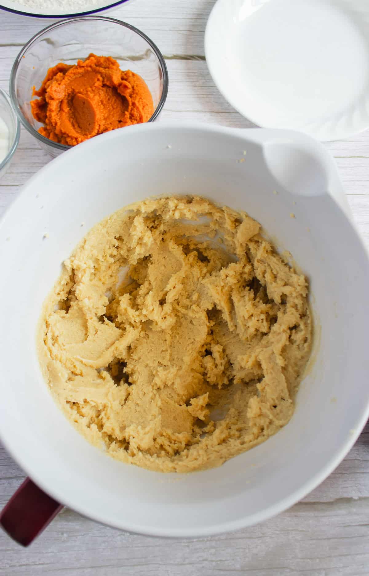 A white mixing bowl with creamed butter and sugar, next to bowls of pumpkin puree and other ingredients—perfect for starting your favorite pumpkin cookies recipe or topping them later with a luscious cream cheese frosting.