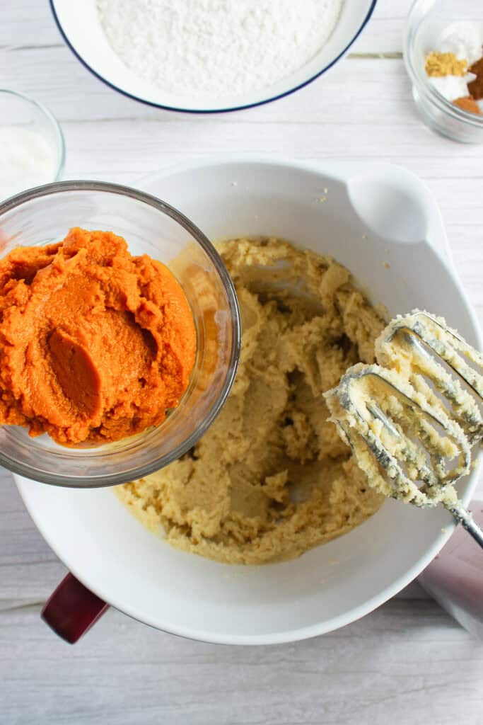Hand holding bowl of pumpkin puree above bowl of cookie dough with electric mixer and ingredients nearby—the perfect start for a delicious pumpkin cookies recipe.