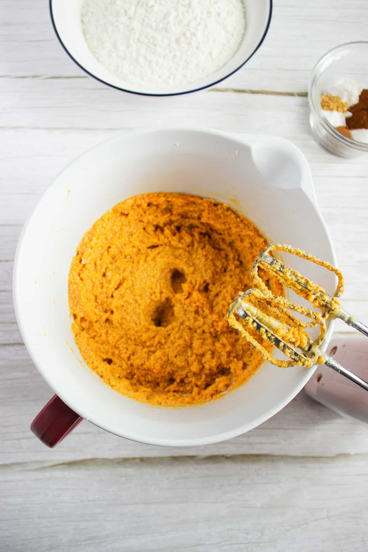 A hand mixer blends orange batter in a white bowl for a delicious pumpkin cookies recipe; flour and spices sit in bowls nearby on a light surface, perfect companions for making Pumpkin cookies topped with creamy cream cheese frosting.