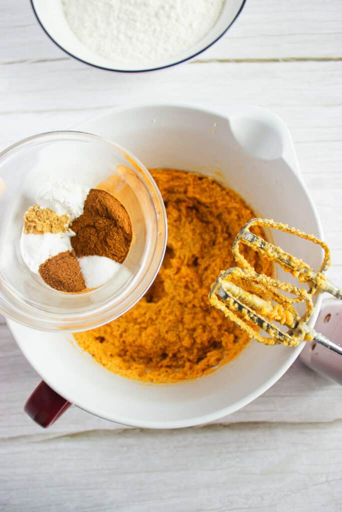 Mixing bowl with orange batter for a delicious Pumpkin Cookies Recipe, beaters, and a glass bowl of spices held above on a white surface—perfect for making Pumpkin Cookies topped with creamy Cream Cheese Frosting.