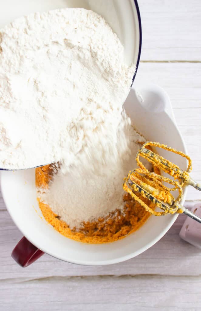 Flour is being poured into a mixing bowl with orange batter for a delicious pumpkin cookies recipe, with a hand mixer ready on the white wooden surface—perfect for making Pumpkin cookies to top with cream cheese frosting.