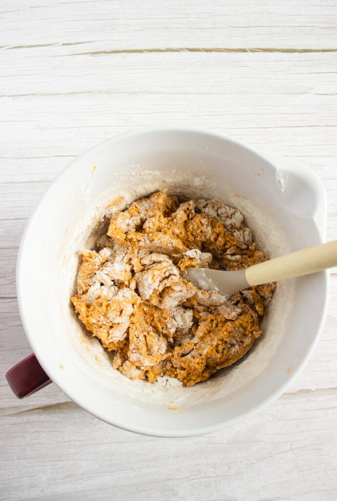 Bowl of partially mixed dough for Pumpkin Cookies with a spatula on a white wooden surface, ready to be transformed into a delicious Pumpkin Cookies Recipe—perfect when topped with creamy Cream Cheese Frosting.