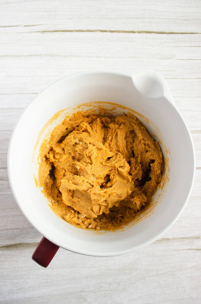 A white mixing bowl filled with thick, light brown pumpkin cookies dough on a white wooden surface, perfect for making a delicious pumpkin cookies recipe topped with cream cheese frosting.