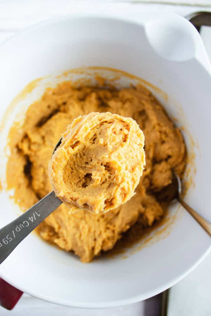 A spoonful of pumpkin cookie dough, perfect for a delicious Pumpkin Cookies recipe, is held over a mixing bowl filled with even more dough and a metal spoon—just waiting to be baked and topped with creamy cream cheese frosting.