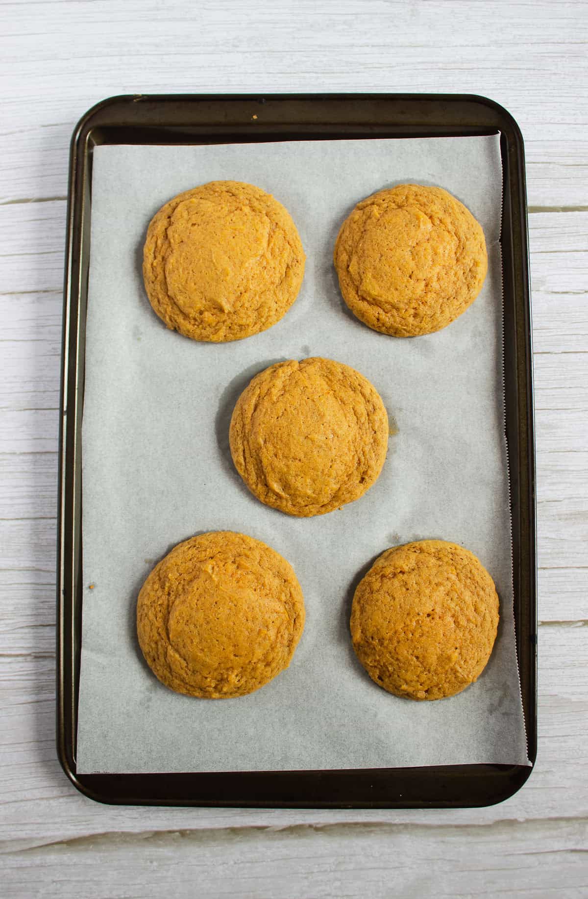 Five round pumpkin cookies on a parchment paper-lined baking tray, on a light wooden surface—a perfect Pumpkin Cookies Recipe ready to be finished with creamy cream cheese frosting.
