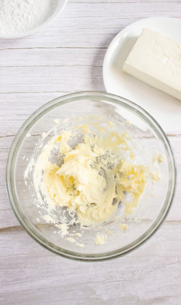 A glass bowl with creamed butter, a stick of butter on a plate, and a bowl of flour on a white surface—essential ingredients for starting your favorite Pumpkin Cookies Recipe or preparing the perfect base for homemade Cream Cheese Frosting.